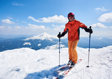 Smiling Sportsman Skier In Helmet And Goggles Standing On Skis Holding Ski Poles In Deep White Snow On Copy Space Background Of Bright Blue Sky And Highland Landscape. Winter Skiing Hobby Concept.