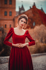 Wall Mural - Beautiful girl in a burgundy red dress walking near  old castle on a background of autumn grape leaves in the park, October. Radomyshl