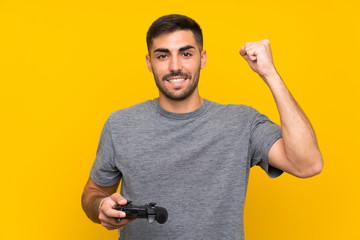 Wall Mural - Young handsome man playing with a video game controller over isolated yellow background celebrating a victory