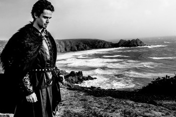 Wall Mural - Knight with leather costume, fur cloak and sword standing in contemplation on cliff top with ocean in background.