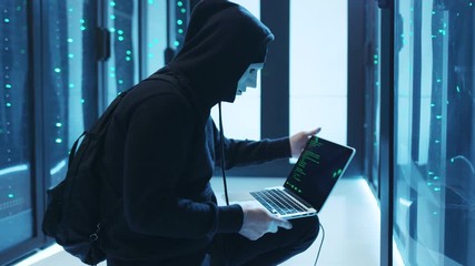 Vengeful hacker in mask programming code on laptop connected to server cabinet. Satisfied spy stealing information breaking data center server running away. Phishing concept.