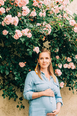 Outdoor portrait of stylish pregnant woman posing in rose garden, wearing denim dress