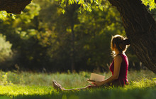 Sunny Portrait Of A Beautiful Girl Sitting On Green Glade Under An Arch Of Tree Branches With Book, Woman Reading Novel On Nature, Concept Hobby And Lifestyle