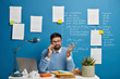 © WHstudio Leushin N - Young male entrepreneur thinks on business solution during telephone conversation, raises hand confusingly, sits at white desk with notepads, crumpled paper and laptop computer, works as manager