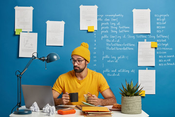 Surprised professional male IT professional freelancer focused in monitor of laptop, tries to improve code of application, drinks coffee and eats sandwich. Skilled geek develops application software
