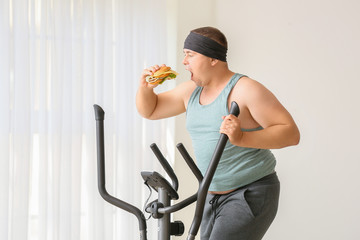 Sticker - Overweight man eating burger while training at home. Weight loss concept