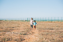 Two Poor Refugee Children Seekers Boy With Toy And Girl Walking Across Hot Desert With State Border Fence On Background Hand In Hand