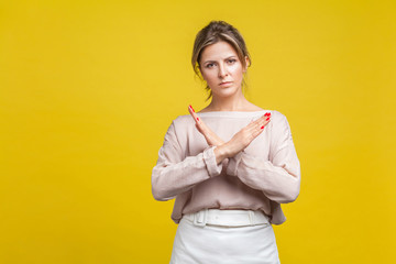 No more, this is the end. Portrait of serious stubborn young woman with fair hair in casual blouse standing showing closed gesture with crossed hands. indoor studio shot isolated on yellow background