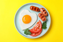 Plate Of Heart Shaped Fried Egg And Sausages On Yellow Background, Top View