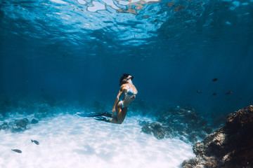 Wall Mural - Freediver girl with fins glides over sandy bottom with fishes in blue ocean