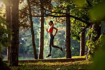 Wall Mural - Side view of slim attractive caucasian brunette in sportswear and with ponytail running on trail in woods. Morning fitness in nature.