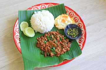 Poster - Stir-fried minced pork with basil leaves served with rice and fried egg on banana leaf on zinc tray over wooden table background.