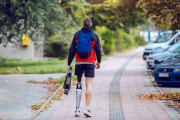 Wall Mural - Rear view of sporty caucasian handicapped man with artificial leg, in sportswear and backpack walking on the street. In hands is artificial leg.