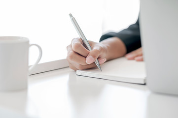 Cropped shot of female hand using pen writing on blank notebook while sitting at office desk.