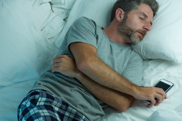 Wall Mural - domestic lifestyle high angle portrait of young attractive and tired man sleeping on bed holding mobile phone in internet and social media addiction and network overuse