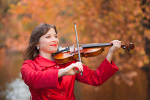 Young Pretty Woman Musician In Red Coat Plays Violin Against Background Of Fall Orange And Yellow Leaves In Autumn Park