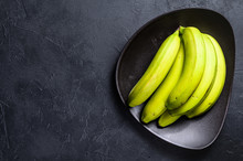 Green Bananas On A Plate. Black Background. Top View. Space For Text. Tropical Fruit