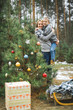 © sofiko14 - Pretty young mother holding her son standing near the decorated Christmas tree in winter forest outdoors. Wooden sledge and present boxes under the tree