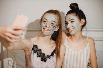 Portrait of two lovely young woman doing selfie laughing while relaxing at home with facial mask on their face. Party for girls relaxing at home.