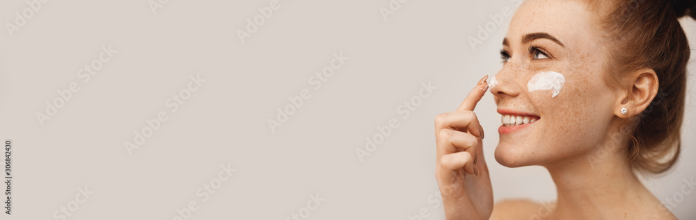 Close up side view portrait of a charming young woman with red hair and freckles applying cream on her face and nose with finger laughing isolated on white wall - obrazy, fototapety, plakaty 