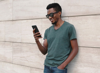 Portrait smiling african man with phone wearing t-shirt, sunglasses on city street over gray wall background