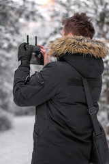 Man holding a drone controller while flying a drone. Black clothing. Shallow depth of field with winter and snowy trees in the background