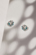 Subject shot of a pair of stud earrings isolated on the beige surface with gray shadows. Each of the earrings is made as a silver flower crown with blue and white gems, pearls and crystals. 