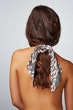 Close-up back shot of a brunette lady with a pink satin hair scarf fixed on the scrunchie with black Paisley print. The shirtless woman with a ponytail is posing on the gray background. 