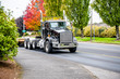 Powerful big rig black semi truck with step down semi trailer and sign for transporting oversize load running on the local road with autumn trees on the side