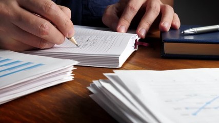 Poster - Hand is writing by pencil in the notepad with financial report and papers.