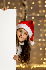 happy little child girl in red santa hat holding white cardboard banner blank with empty space on background with lights