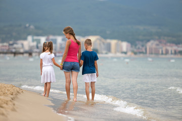 Mother and two children son and daughter walking together on sand beach in sea water in summer with bare feet in warm ocean waves.