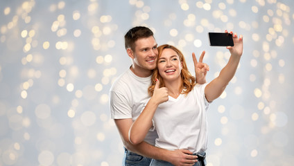 Poster - relationships, technology and people concept - happy couple in white t-shirts taking selfie smartphone and showing peace by over holidays lights background