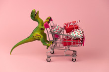 Funny Green Dinosaur Toy With Shopping Cart Full Of Present Boxes On A Soft Pink Background