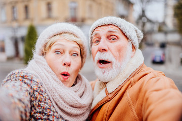 Traveling senior couple wearing warm clothes taking a selfie together kissing against the background of attractions of old city street in cold winter outdoor.