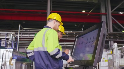 Two diverse male engineers in uniform and hard hats use computer LCD touch screen for programming CNC machine. Work process on heavy industry plant or factory. Employees and high tech