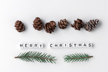 Creative Layout Of Words Merry Christmas Made Of Wooden Cubes With Letters On White Paper Background, New Year Minimalistic Concept With Fir Cones And Green Branches With Needles