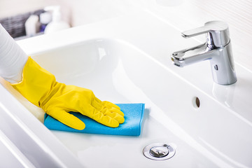 Woman maid or charwoman cleaning modern new basin in bathroom, water tap clean using yellow gloves and blue cloth.