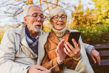 senior couple sitting in public park using smart device to shop online. cheerful seniors using moder