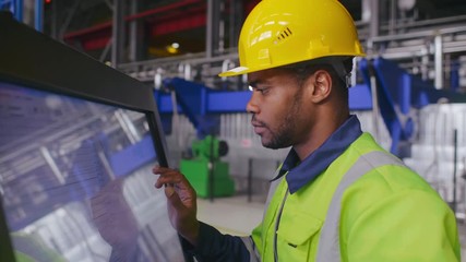 Black male engineer in uniform uses computer LCD touch screen for programming CNC machine or assembling components on conveyor. Work process on heavy industry plant or factory