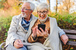 © lordn - Senior couple sitting in public park using smart device to shop online. Cheerful seniors using modern technology.