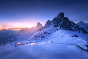 Wall Mural - Snowy mountains and blurred car headlights on the winding road at night in winter. Beautiful landscape with snow covered rocks, house, mountain roadway, blue starry sky at sunset in Dolomites, Italy