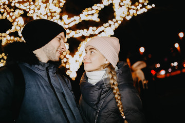 Wall Mural - Loving young couple in winter clothes on a background of Christmas lights. New Year's date.