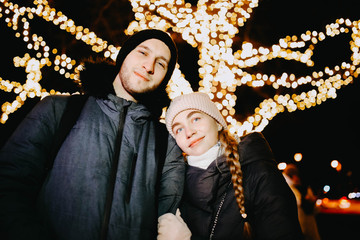 Wall Mural - Loving young couple in winter clothes on a background of Christmas lights. New Year's date.