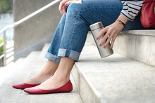 Crop Shot Of An Attractive Woman Wearing Jeans With Stylish Ruby Red Flat Shoes Sitting On A Stairs, Her Hand Grab A Reusable Isolated Water Bottle. Plastic-free, No Straw, Eco, Zero Waste Concept.