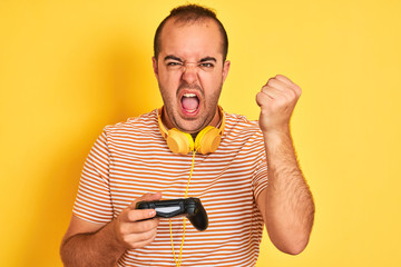 Sticker - Young gamer man playing video game using headphones over isolated yellow background annoyed and frustrated shouting with anger, crazy and yelling with raised hand, anger concept