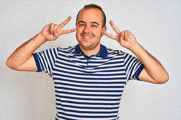 Young man wearing casual striped polo standing over isolated white background Doing peace symbol with fingers over face, smiling cheerful showing victory