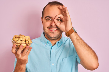 Canvas Print - Young man holding bowl with peanuts standing over isolated pink background with happy face smiling doing ok sign with hand on eye looking through fingers