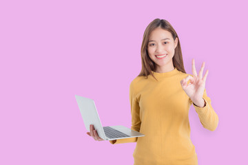 Wall Mural - Beautiful young asian woman holding notebook computer and showing OK sign with smiles over pink background