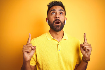 Canvas Print - Young indian man listening to music using earphones standing over isolated yellow background amazed and surprised looking up and pointing with fingers and raised arms.